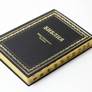 Può includere: Un libro con copertina rigida nera con bordi dorati e scritte dorate sulla copertina. La copertina del libro presenta un bordo dorato decorativo. Il testo sulla copertina è in una lingua straniera.