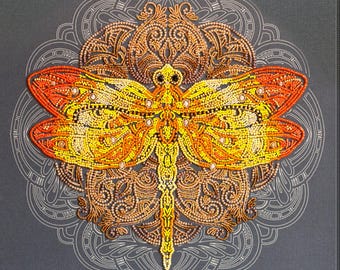 May include: A detailed, beaded dragonfly design in shades of yellow, orange, and gold, set against a gray background. The dragonfly is the focal point, with intricate patterns and embellishments. The artwork is framed by a decorative, circular design.