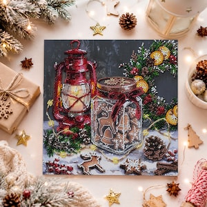 May include: A square diamond painting kit featuring a red lantern next to a glass jar filled with gingerbread cookies shaped like reindeer, bells, and stars. The scene includes a pine wreath with orange slices, pinecones, and red berries.