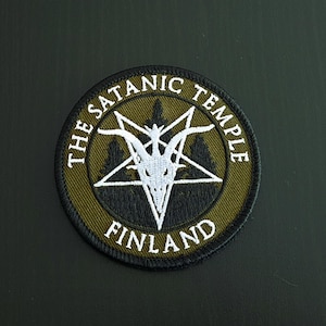 Może przedstawiać: Okrągła naszywka z napisem "THE SATANIC TEMPLE FINLAND" w białych literach. Naszywka przedstawia biały pentagram z głową kozła pośrodku, na zielono-czarnym tle.