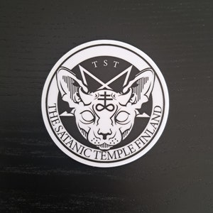 May include: A white circular sticker featuring a black and white illustration of a cat with horns and a pentagram with an infinity symbol inside. The text "The Satanic Temple Finland" is written around the edge of the sticker.