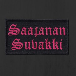 Könnte beinhalten: Schwarzer gestickter Aufnäher mit den Worten "Saatanan Suvakki" in pinker gotischer Schrift.