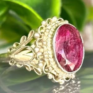 May include: A silver ring with an oval, deep red gemstone. The ring has an ornate, filigree design around the stone and on the band. The background features green foliage, creating a contrast with the ring's metallic and gemstone colors.