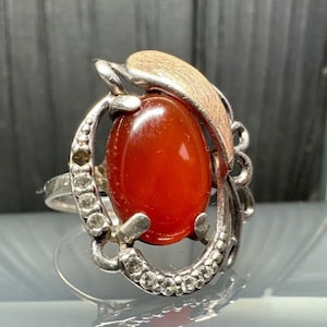 May include: A silver ring featuring an oval, reddish-orange gemstone. The stone is set in a decorative silver setting with small, clear accent stones. A gold-toned leaf-like element adorns the top of the ring, adding a unique touch.
