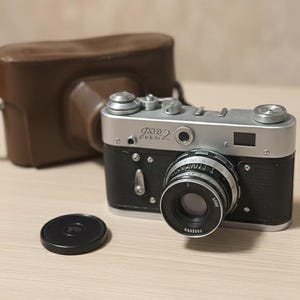 Puede incluir: Una cámara vintage plateada y negra con una tapa de lente y una funda de cuero marrón. La cámara tiene el texto "FED 2" en la parte superior. La lente es negra y plateada. La funda está detrás de la cámara.