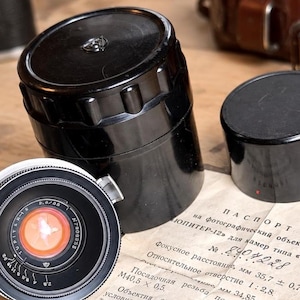 May include: A black Jupiter-12 camera lens with a silver rim, two black cylindrical storage cases, and a vintage paper passport document featuring technical specifications in Cyrillic text, including a focal length of 35.7 mm and aperture 1:2.8.