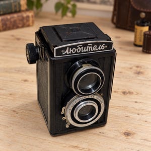 May include: A black twin-lens reflex camera featuring a top-mounted viewfinder and a front plate with the Cyrillic text "Любитель". The lens assembly includes silver metal rings with markings indicating 7.5cm, 1:4.5, and other technical specifications.