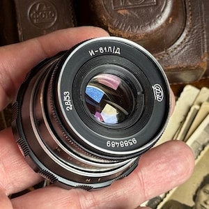 Peut inclure: Un objectif d'appareil photo vintage avec un boîtier en métal noir et argenté. L'objectif a un élément en verre réfléchissant et est marqué de chiffres et de texte cyrillique. L'objectif est tenu dans une main, avec un étui en cuir visible en arrière-plan.