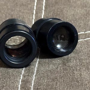 Puede incluir: Dos lentes &oacute;pticas negras y cil&iacute;ndricas con una abertura circular. Las lentes est&aacute;n colocadas una al lado de la otra sobre una superficie marr&oacute;n texturizada. Las lentes parecen estar hechas de un material duradero.