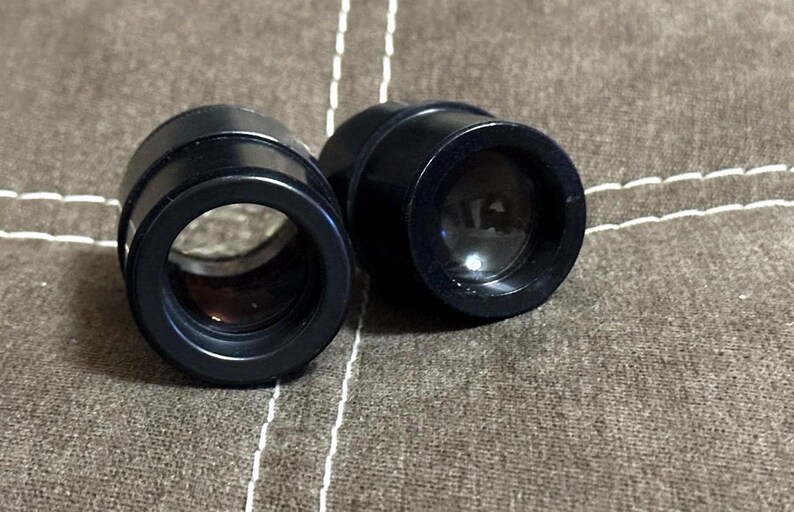 Puede incluir: Dos lentes &oacute;pticas negras y cil&iacute;ndricas con una abertura circular. Las lentes est&aacute;n colocadas una al lado de la otra sobre una superficie marr&oacute;n texturizada. Las lentes parecen estar hechas de un material duradero.