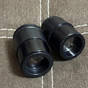 Puede incluir: Dos lentes cil&iacute;ndricas negras con interior reflectante. Las lentes tienen una parte superior estriada y un exterior liso y brillante. Se encuentran sobre una superficie de tela marr&oacute;n texturizada.