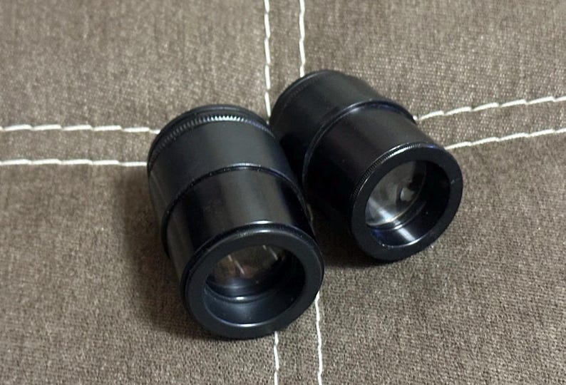 Puede incluir: Dos lentes cil&iacute;ndricas negras con interior reflectante. Las lentes tienen una parte superior estriada y un exterior liso y brillante. Se encuentran sobre una superficie de tela marr&oacute;n texturizada.