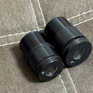 Puede incluir: Dos oculares de microscopio negros con anillos de ajuste moleteados. Un ocular est&aacute; marcado con "8x". Los oculares est&aacute;n sobre una superficie de tela marr&oacute;n texturizada.