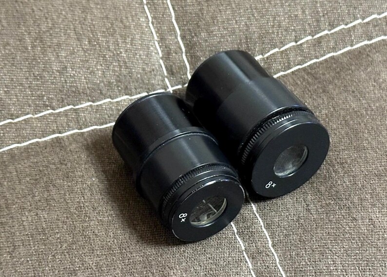 Puede incluir: Dos oculares de microscopio negros con anillos de ajuste moleteados. Un ocular est&aacute; marcado con "8x". Los oculares est&aacute;n sobre una superficie de tela marr&oacute;n texturizada.