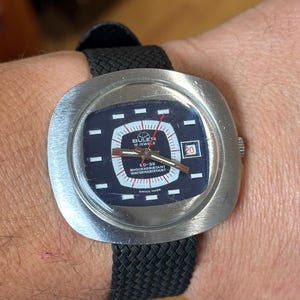 Könnte beinhalten: Eine Vintage Buler CD-35 Swiss Made Uhr mit einem schwarzen gewebten Armband. Das Zifferblatt ist dunkelblau mit weißen und roten Akzenten, mit Datumsanzeige auf 3 Uhr. Die Uhr hat ein Edelstahlgehäuse.