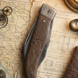 Pode incluir: Uma faca dobrável vintage com cabo castanho e lâmina prateada, repousando sobre um mapa e papel envelhecido. O cabo apresenta um padrão decorativo. A faca é uma ferramenta clássica.