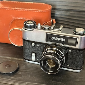 Puede incluir: Una cámara de película de 35 mm plateada y negra con una funda de cuero. La cámara tiene un objetivo con una tapa negra y el texto "ФЭД 5B" en la parte superior.