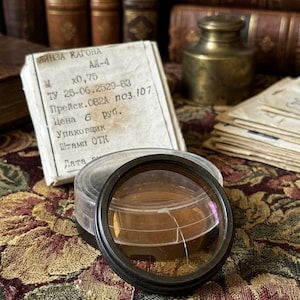 May include: A vintage camera lens filter with a black rim and a clear plastic case. The filter has a yellow tint and a crack. A box with Russian text is behind the lens, along with antique books and a brass container.