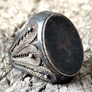 Peut inclure: Bague argentée avec une pierre ovale gris foncé. La bague présente des détails de filigrane complexes sur les côtés et une bordure en forme de corde autour de la pierre. Posée sur une surface en bois texturée.