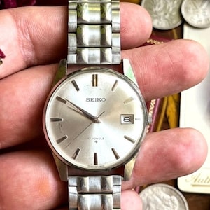 May include: A silver-toned Seiko wristwatch featuring a round metallic dial with baton hour markers, silver hands, and a date window at the three o'clock position. The watch includes a 17 jewels movement and a matching silver-toned metal link bracelet.