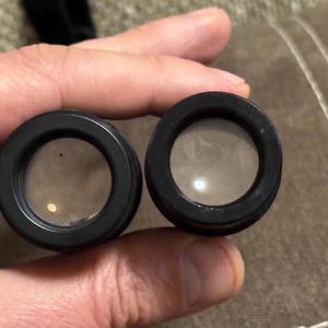 Puede incluir: Dos lentes negras y circulares con vidrio transparente, posiblemente para un telescopio o binoculares. Las lentes se sostienen en una mano, con un fondo neutro. Las lentes tienen un acabado oscuro y mate.