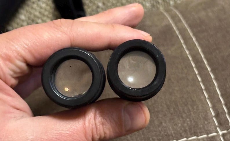 Puede incluir: Dos lentes negras y circulares con vidrio transparente, posiblemente para un telescopio o binoculares. Las lentes se sostienen en una mano, con un fondo neutro. Las lentes tienen un acabado oscuro y mate.