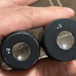 Puede incluir: Dos lentes cil&iacute;ndricas negras con vidrio transparente, cada una marcada con "x8" en blanco. Las lentes se sostienen en una mano, lo que sugiere que son parte de un dispositivo de aumento o un instrumento &oacute;ptico.