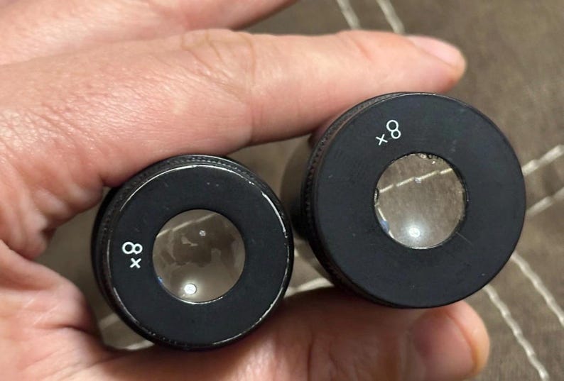 Puede incluir: Dos lentes cil&iacute;ndricas negras con vidrio transparente, cada una marcada con "x8" en blanco. Las lentes se sostienen en una mano, lo que sugiere que son parte de un dispositivo de aumento o un instrumento &oacute;ptico.