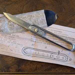 May include: A silver-toned folding pocket knife with a metallic skeletonized handle and a pointed blade. The knife rests on a light tan paper document featuring technical diagrams and Cyrillic text, accompanied by a textured beige protective sleeve.