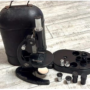 May include: A vintage black microscope with a silver eyepiece and a round stage. The microscope is accompanied by a black carrying case and a tray with several lenses. The set is designed for scientific study.