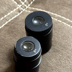Puede incluir: Dos lupas cil&iacute;ndricas negras con aumento de 8x. Cada lupa tiene una lente y un anillo de enfoque moleteado. Las lupas est&aacute;n sobre una superficie marr&oacute;n texturizada.