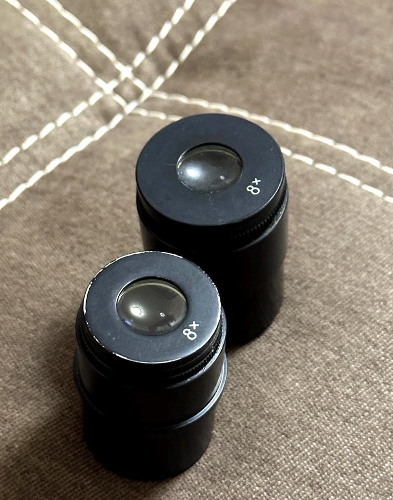 Puede incluir: Dos lupas cil&iacute;ndricas negras con aumento de 8x. Cada lupa tiene una lente y un anillo de enfoque moleteado. Las lupas est&aacute;n sobre una superficie marr&oacute;n texturizada.