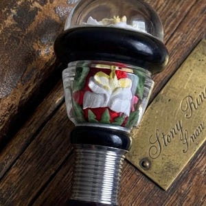 Peut inclure: Une poignée décorative avec un dôme en verre transparent, un anneau noir et une section centrale affichant une fleur blanche et jaune avec des feuilles vertes sur fond rouge. La base comprend un col en métal argenté strié et un manche sombre.