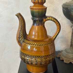May include: A ceramic pitcher with a spout and handle, in brown and orange glaze with decorative patterns. The pitcher has a lid with a green top, likely for serving beverages. The design is vintage and decorative. The pitcher is approximately 25 cm tall.