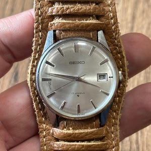 May include: A vintage Seiko wristwatch with a silver face and a brown leather cuff-style band. The watch face displays the brand name, Seiko, and the words "17 Jewels". The watch has a date window at the 3 o'clock position.