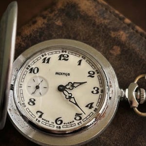 Può includere: Un orologio da tasca color argento con quadrante color crema, numeri arabi neri e un quadrante secondario. Il quadrante riporta la scritta Molnija e Made in Russia. L'orologio presenta lancette nere decorate e una cassa in metallo con corona.