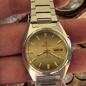 May include: A silver-toned Orient wristwatch with a metallic gold dial, gold-colored hour markers, and gold hands. The watch features a day-date window at the three o'clock position, the Orient logo, and the text AAA on the dial. It has a metal link band.