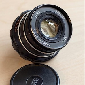 May include: A black Industar-61 L/D camera lens with a 2.8/55 aperture and focal length specification. The lens features silver metallic rings, Cyrillic text, and a serial number 9322263. A separate black circular lens cap is positioned below.