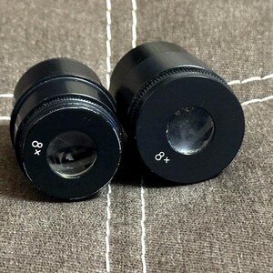 Puede incluir: Dos oculares de microscopio negros con la marca "8x" en blanco. Los oculares son cil&iacute;ndricos con una lente circular en el extremo. El fondo es una tela texturizada de tono neutro.