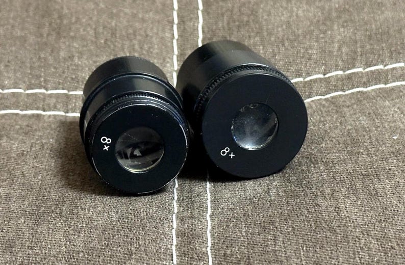 Puede incluir: Dos oculares de microscopio negros con la marca "8x" en blanco. Los oculares son cil&iacute;ndricos con una lente circular en el extremo. El fondo es una tela texturizada de tono neutro.