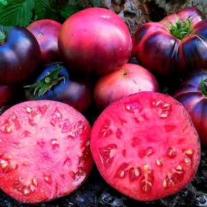 May include: A selection of ripe heirloom tomatoes in shades of purple, red, and pink. One tomato is sliced open, showing the juicy flesh. The tomatoes are displayed on a dark, textured surface.