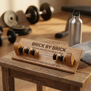 Op de afbeelding: Een houten workout-tracker in abacus-stijl met zwarte en naturel houten kralen op een metalen staaf. De basis is gegraveerd met de tekst BRICK BY BRICK, One rep, one set, one day at a time, en 10 beads moved -> Watch the strength grow.
