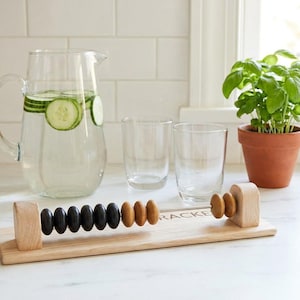 Op de afbeelding: Een houten abacus-achtige teller met een lichte houten basis en twee rechtopstaande steunen. Het heeft een metalen staaf met tien zwarte kralen en vijf natuurlijke houtkleurige kralen. De basis heeft de gedeeltelijke tekst RACKER in het zwart.