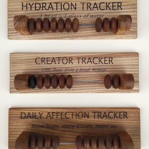 May include: Three rectangular wooden boards with abacus-style sliding beads. The top board reads HYDRATION TRACKER 1 bead = 1 glass of water. The middle board reads CREATOR TRACKER Film. Post. Slide a bead. Repeat. The bottom board reads DAILY AFFECTION TRACKER More hugs, more kisses, more us.