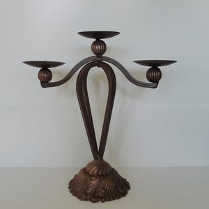 Peut inclure: Un candélabre en métal brun foncé doté de trois coupelles circulaires pour bougies, soutenues par des accents sphériques nervurés. La tige centrale se compose de deux barres métalliques courbées formant une boucle allongée sur une base festonnée.