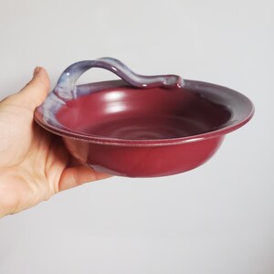 Peut inclure: Un bol en céramique rond avec une glaçure marron profond et une finition lisse et brillante. Le bol présente une anse unique et incurvée s'étendant depuis le bord, rehaussée d'une glaçure bleue et blanche contrastant avec le corps rouge foncé.