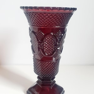 May include: A deep red glass vase featuring a flared rim and a textured diamond pattern across the body. The vessel includes raised oval motifs and sits on a sturdy, hexagonal pedestal base, showcasing a translucent finish and intricate detailing.