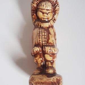 May include: A ceramic figurine of a person wearing a hooded parka with a textured fur-lined trim. The figure is rendered in a two-tone colour scheme of cream and dark brown, standing on a rectangular base with a smooth, glossy finish throughout.