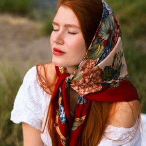 Peut inclure: Un foulard en soie rouge à motifs floraux drapé sur la tête et les épaules. Le foulard présente un fond crème avec de grandes fleurs colorées et une bordure rouge foncé. Le foulard est associé à un haut blanc à épaules dénudées.