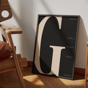 Peut inclure: Une affiche encadrée noire et beige avec une grande lettre "G". L'œuvre est appuyée contre un mur blanc et est éclairée par la lumière naturelle. L'impression contient du texte en haut et en bas.